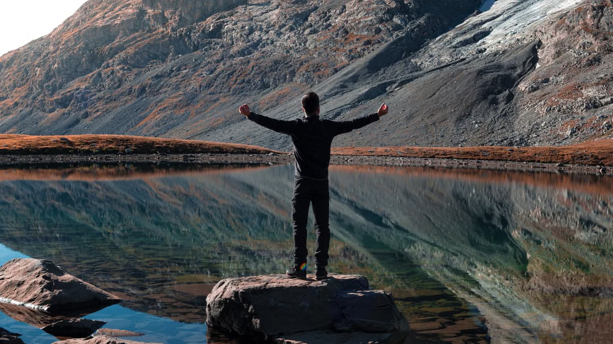A man stands with outstretched arms by a mountain lake, reflecting rocky slopes in Auvergne-Rhône-Alpes.